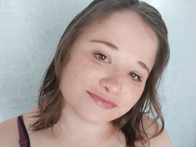 Annoying has wet brown hair, light skin, freckles, and pink lipstick, wearing a dark top, with a subtle smile, showing small tits and brown eyes.