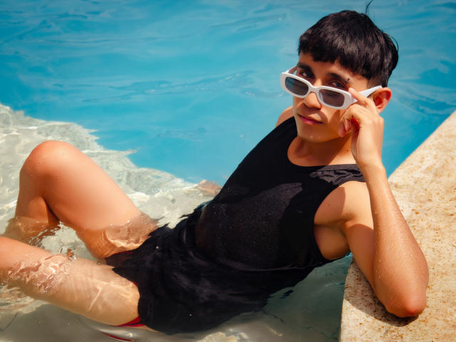 ThaylerJones, a cute young dude with dark hair, wearing white sunglasses and a wet black sleeveless shirt, lounges in a blue pool, his red shorts partially visible.