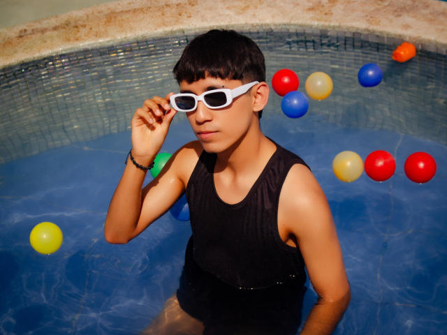 ThaylerJones, young Asian dude, in blue water, wearing white-framed glasses, black wet tank top, adjusting glasses, with colorful balls in the background, short black hair, tan skin.