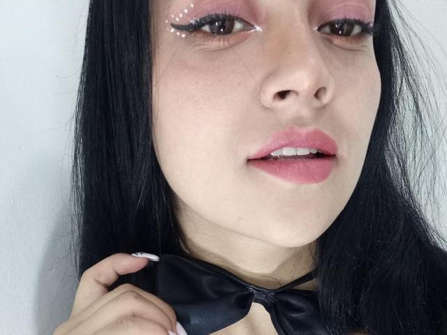 SexyEuphorua with pink eyeshadow, white eyeliner dots, black hair, pink lips, holding a black bowtie, showing a slight smile, and a hand with white nails in the shot.