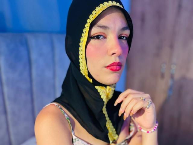 AliceKayleigh with a black and yellow headscarf, pink lipstick, and a ring on her right hand, showing off her painted nails, sitting with a slightly flushed face.