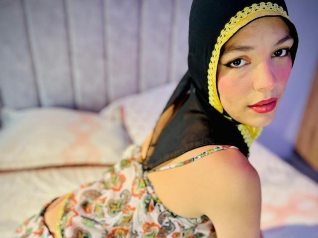 AliceKayleigh with a black hijab and yellow lace, looking over her shoulder, wearing a floral dress, showing her bare shoulders and red lipstick, sitting on a bed.