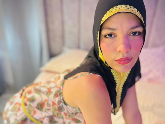 AliceKayleigh with a black and yellow headscarf, wearing a floral dress, leaning forward on a bed, showing her red lipstick and cute smile, with her boobs slightly visible.