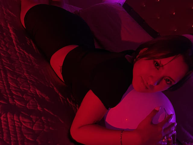SkarlethStiv lying on a bed, wearing a black top and shorts, holding a white ball, with red and purple lighting, looking at the camera, showing her tits.