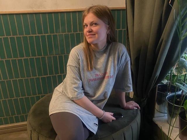 AuroraAngel sits on a green cushion, wearing a gray shirt dress and black tights, with a small butt and medium tits, looking to the side with a smile.