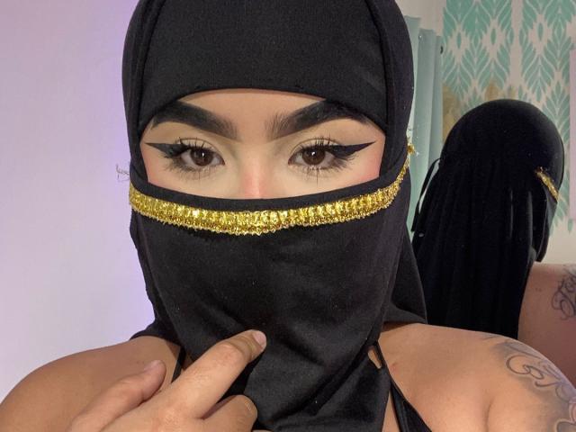 DaliilaAli, con un pañuelo negro adornado con detalles dorados y un delineador de ojos llamativo, mostrando el dedo hacia la boca. Se puede ver un tatuaje en el hombro derecho. El reflejo en el espejo muestra la parte posterior del pañuelo..