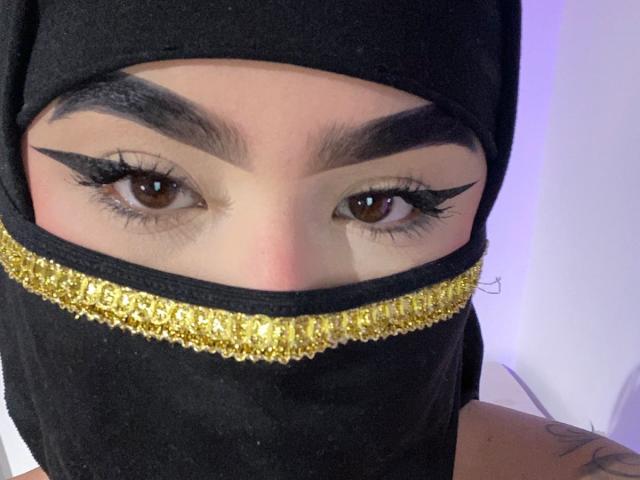 DaliilaAli with dark eyebrows, bold eyeliner, brown eyes, black headscarf, gold embroidered band, and tattoo on visible arm, showing only face and upper neck.