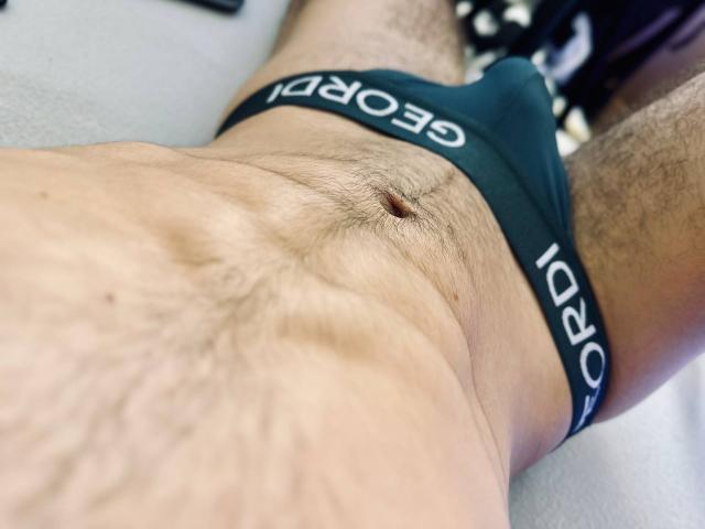 Jakes Magnus' unterer Körper, bekleidet mit dunkelgrünen Boxershorts, zeigt einen behaarten Bauch, die Bauchnabe und einen Teil seines Penis, wobei auch Haare an den Oberschenkeln sichtbar sind. Seine Haut ist hell und leicht strukturiert..