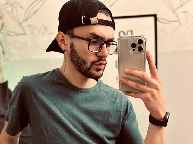 Jake Magnus che si fa un selfie, indossa un berretto nero, occhiali, una maglietta verde, un orologio da polso e ha la barba, tiene in mano un iPhone argentato, ha un tatuaggio sull'orecchio e un'espressione seria..