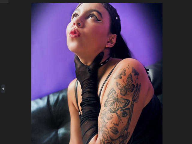 NenaBlack with pouty lips, black gloves, and butterfly tattoo on her right arm, her dark hair slicked back, purple background, black leather couch, and a sexy, sultry expression.