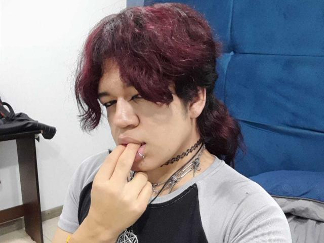 Spectrum69 with dark red, messy hair, gray and black shirt, finger in mouth, choker necklace, tattoos on neck, blue chair in background, looking down with a pouty expression.