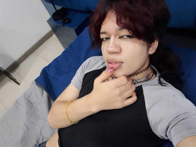 Spectrum69 with dark red hair, grey and black raglan shirt, chomping on finger, black choker, tattoo on neck, yellow bracelet, lying on blue and grey bed.