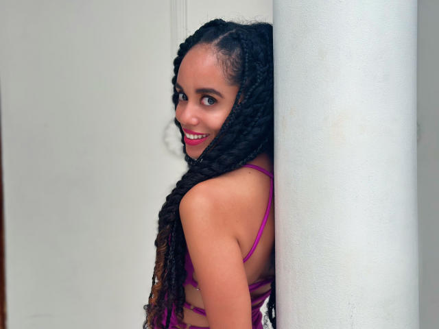 TerryGreen with long black braids, purple bikini, smiling, standing behind a white pillar, showing her back and looking over her shoulder with red lipstick.