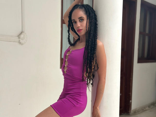 TerryGreen with long black dreadlocks, wearing a tight purple dress, leaning against a white pillar, showing her small tits and slim ass. Her arm is raised, looking at the camera.