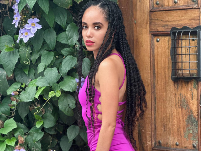 TerryGreen with long, dark braided hair wearing a tight, pink dress, standing in front of a wooden door with a metal handle. Her ass is visible.