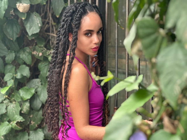 TerryGreen with long, dark braided hair, wearing a tight, purple dress, standing near green foliage, looking back with red lipstick, showing a cute, sexy, brown-skinned face.