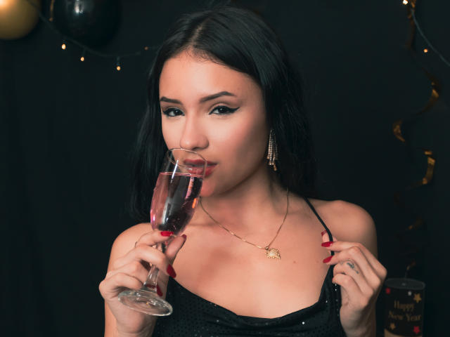 LizzeJoy with dark hair, drinking pink wine, holding glass to mouth, black dress, red nails, gold necklace, earrings, and small tattoo on right hand, against black background.