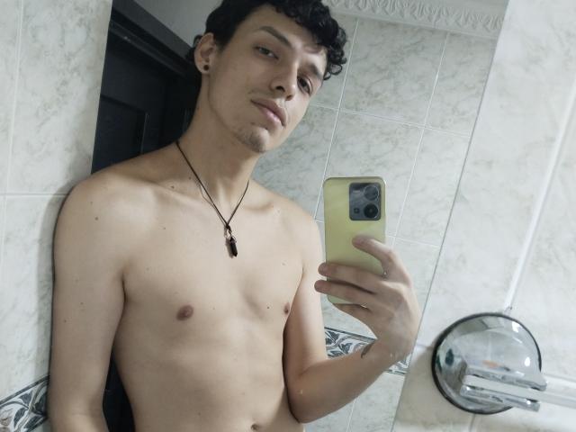 LinoRayn, shirtless, with curly black hair, holding a yellow phone, wearing a necklace with a black pendant, taking a mirror selfie, showing a slight smile, in a tiled bathroom.