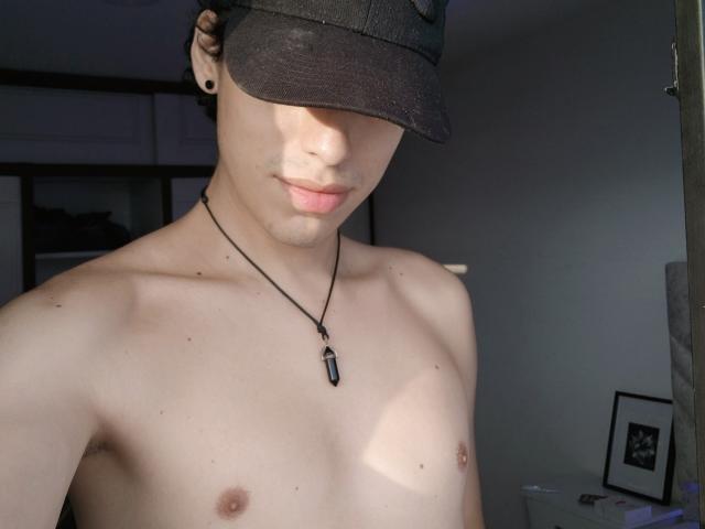 LinoRayn is shirtless, wearing a black cap, necklace with a black pendant, and black ear gauges, with light skin and small nipples, looking down with a slight smile.