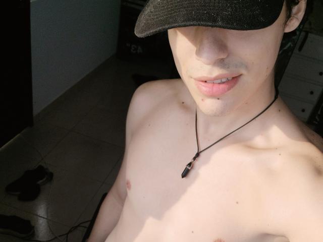 LinoRayn's shirtless upper body, wearing a black cap, necklace with a pendant, and light skin with visible nipples, subtle lip piercing, and a slight smile.