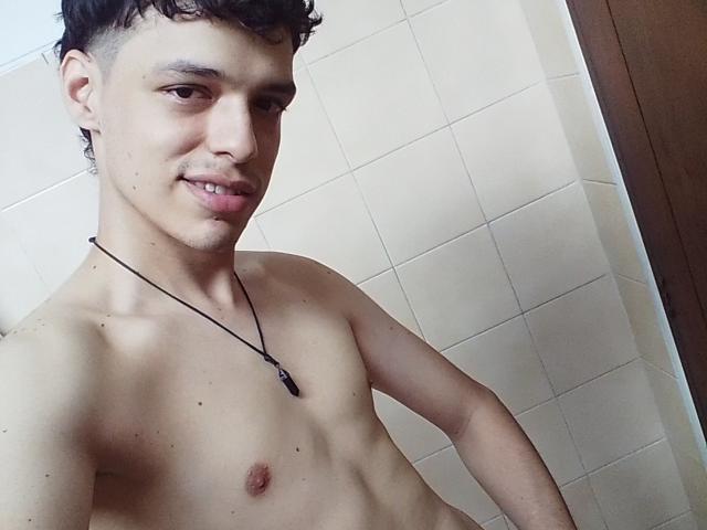 LinoRayn, shirtless with short curly black hair, light skin, small chest, necklace, slight smile, standing against tiled wall, showing a small, slightly raised nipple.