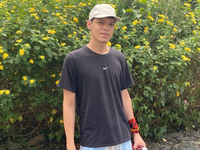 LinoRayn, wearing a black Nike shirt, white cap, and blue shorts, stands in front of yellow flowers. He's got a red wristband and a slight smile.