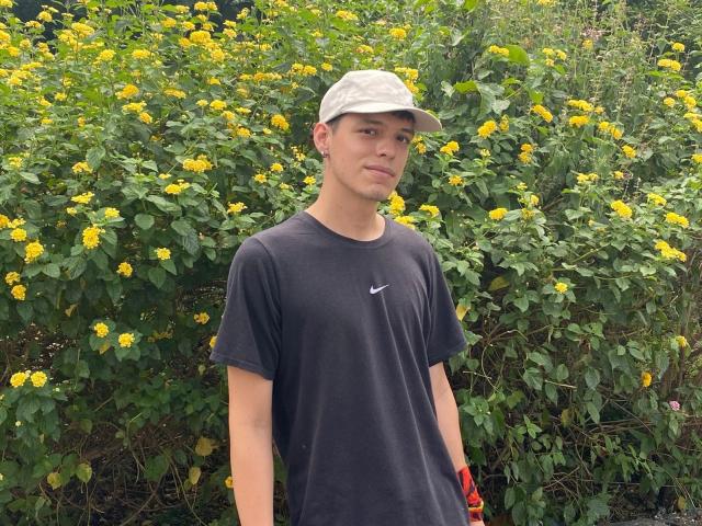 LinoRayn stands in front of a bush with yellow flowers, wearing a grey cap, black Nike t-shirt, and red wristband, with a slightly bored expression.