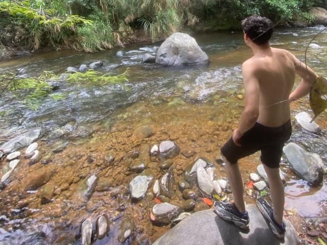 LinoRayn stands shirtless on a rock, facing a clear creek with visible stones and small orange fish. He's wearing brown shorts and black sneakers. His back is to the camera.