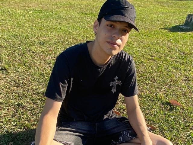 LinoRayn, sitting on grass, wearing a black cap, black t-shirt with a cross, and denim shorts, has a slightly dirty face, and a small bruise on his left cheek.
