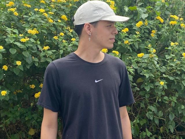 LinoRayn with short black hair, white cap, black Nike t-shirt, small earrings, standing in front of yellow flowers, green leaves, looking to the right, showing a cute profile.