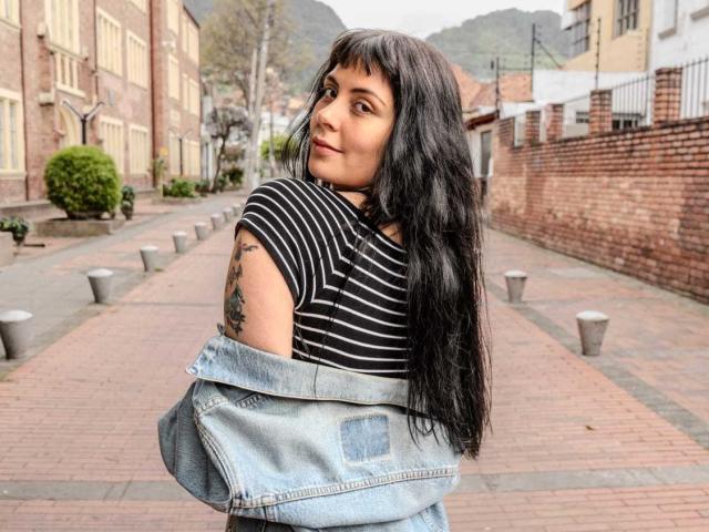 GiaAmatto with long black hair, striped shirt, and blue denim jacket, looking back, showing tattoo on right arm, standing on brick walkway.
