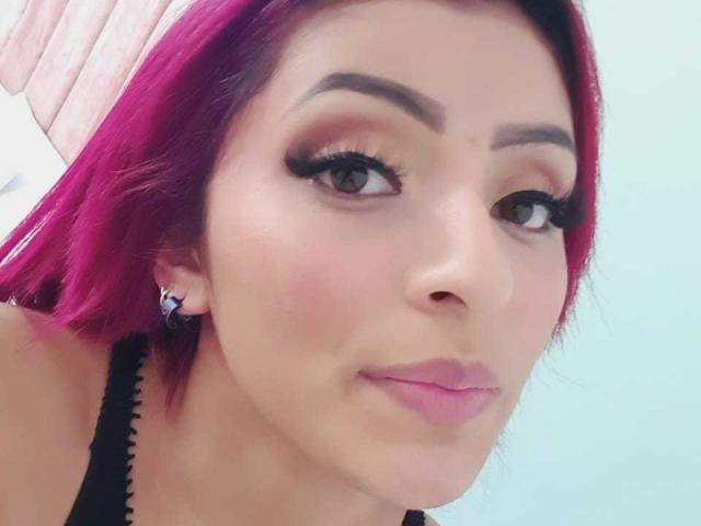 LizBonnet with pink hair, thick eyebrows, long black eyelashes, and pink lipstick, pouting her glossy lips. She's wearing black strap earrings and a black top.