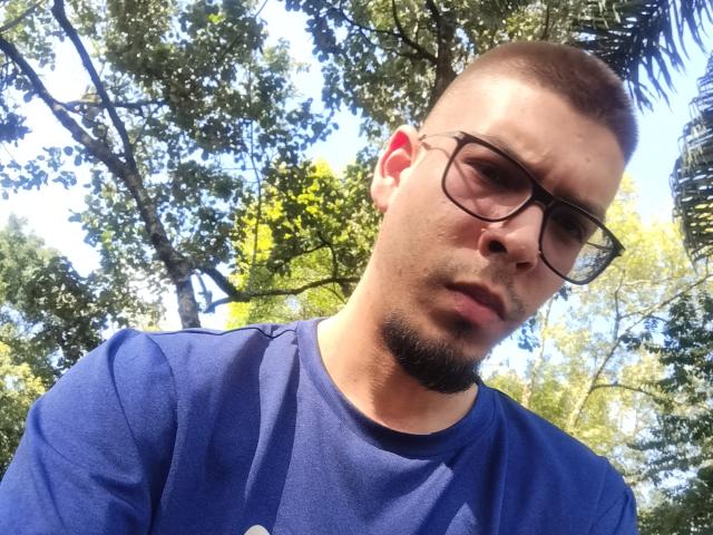 GolFather has a short brown haircut, glasses, and a goatee. He's wearing a blue shirt, looking slightly down with a serious expression. Sunlight filters through green trees.