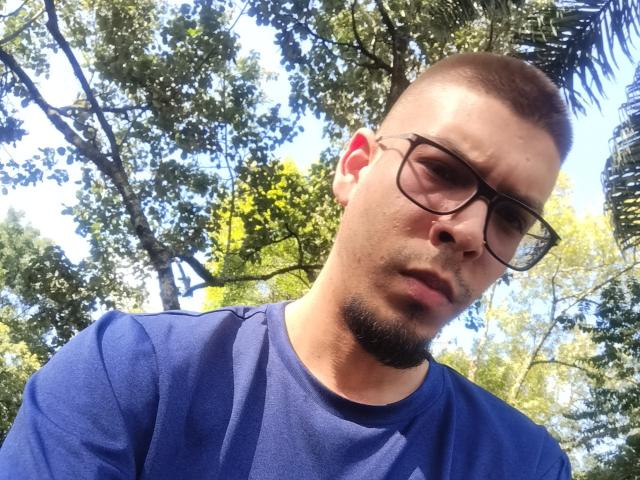 GolFather has a serious face, short brown hair, beard, glasses, and is wearing a blue shirt in a sunny outdoor setting. His nose is slightly upturned.