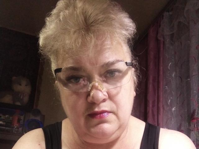 OneDamaX has curly gray hair, glasses, a nose ring, and pink lipstick. She's wearing a black tank top, looking directly at the camera with a serious expression.