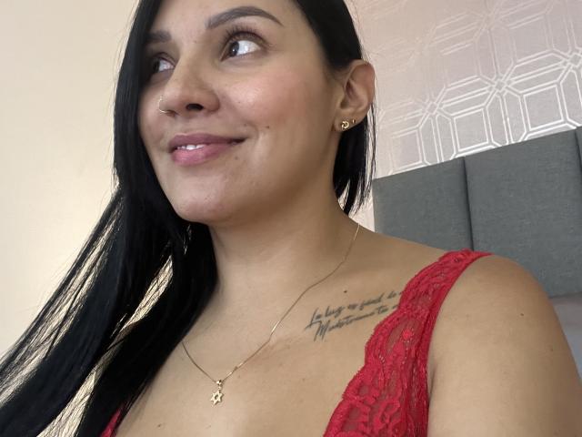 YessiJaymes, com cabelos pretos longos, pele clara e pequenos brincos de ouro, sorrindo levemente, vestindo uma blusa vermelha de renda, mostrando uma tatuagem em seu ombro direito..