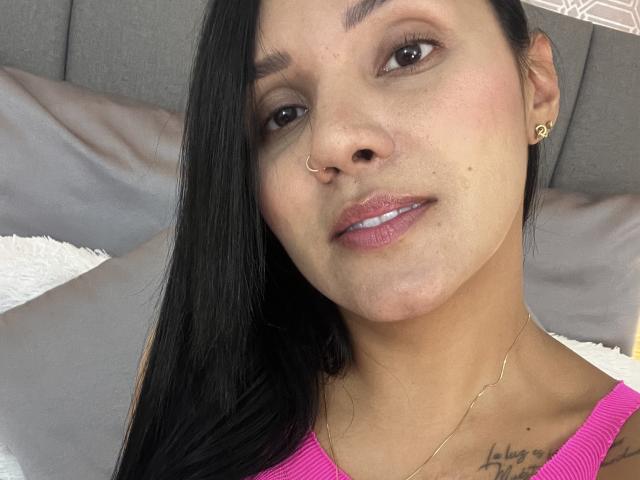 YessiJaymes has light brown skin, black hair, a nose ring, small gold earrings, pink lipstick, and a tattoo on her right chest. She's wearing a pink top.