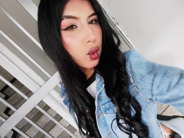 AnnyyHot, avec ses longs cheveux noirs, ses lèvres roses et pulpeuses, et son trait d'eye-liner épais, porte une veste en jean bleu et regarde l'appareil photo avec une bouche légèrement entrouverte..