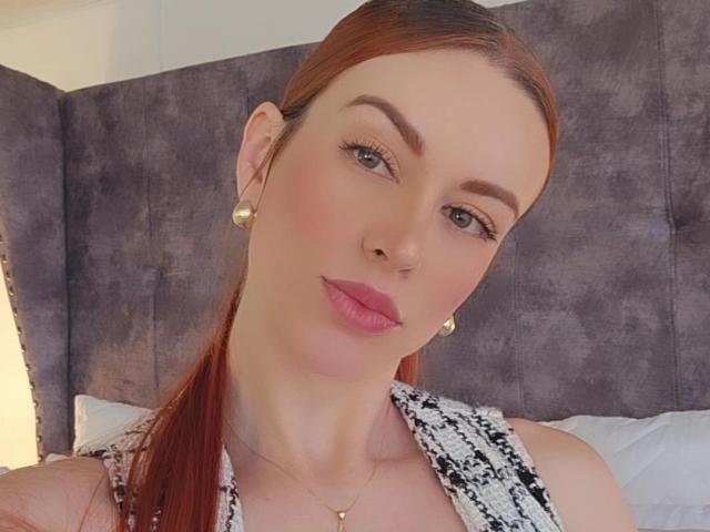 Leared has red hair, pink lips, and is wearing gold earrings. She's looking at the camera with a slight smile, showing off her tits and a black-and-white patterned top.
