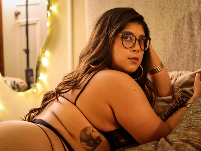 IsabelArt with long brown hair, glasses, and hoop earrings, lying on a bed in black lingerie, showing her butt and a tattoo on her side, looking back.