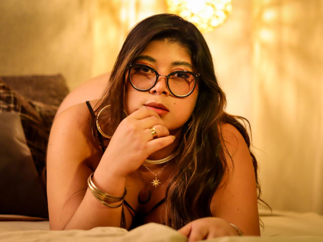 IsabelArt with glasses, brown hair, and a necklace, lying on a bed, hand near her mouth, showing her tits, wearing a ring and bracelets, in a warm, golden light.
