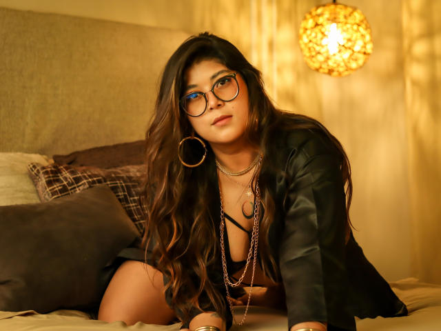 IsabelArt with long brown hair, blue eyeshadow, glasses, black shirt, necklace, hoop earrings, kneeling on bed, looking at camera, soft yellow lighting, warm, cozy, sexy.