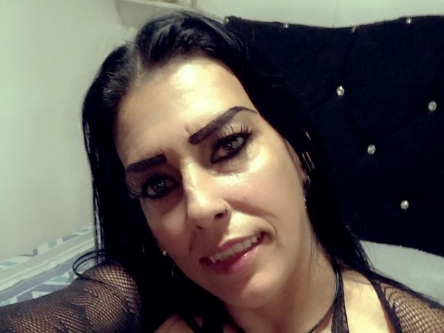 LagaGomez, con i capelli neri, indossa una maglia a rete, fa l'occhiolino con un leggero sorriso, mostrando un piccolo taglio sulla guancia e un trucco per gli occhi molto marcato. Il suo sedere è parzialmente visibile..