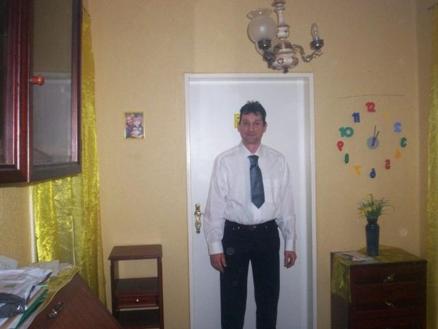 Mario41-hot stands in a room, wearing a white shirt, black tie, and black pants, with a slightly goofy expression, in front of a white door.