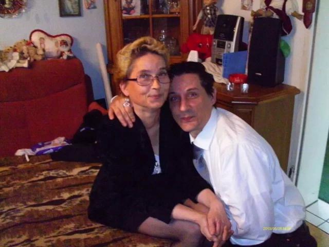 Mario41-hot with short blonde hair, glasses, and black dress, sits on brown table, arm around Mario41-hot in white shirt, black tie, and dark hair, both smiling.