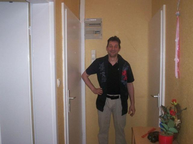 Mario41-hot, standing in a hallway, wearing a black shirt with red emblem, beige pants, and a black belt, smiling with hands on hips, short brown hair, casual look.