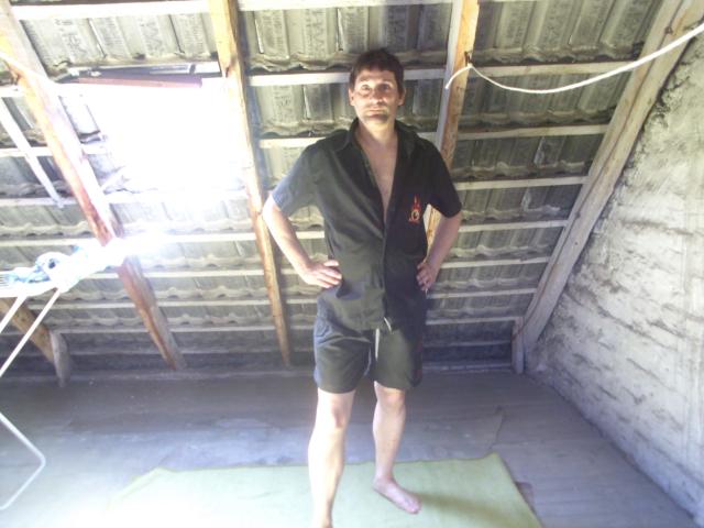 Mario41-hot stands barefoot in a rustic shed, wearing an unbuttoned black shirt and green shorts, with hands on hips, showing a slight smile, and short brown hair.