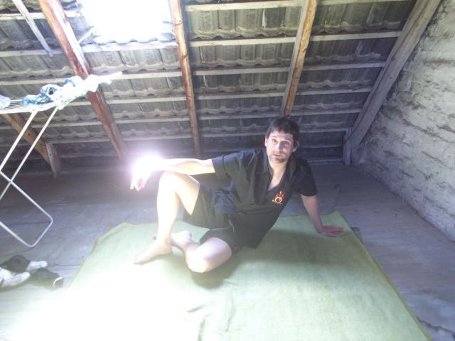Mario41-hot, a nerdy dude with short brown hair, wearing a black shirt and shorts, sits on a green mat in a dim attic, legs crossed, looking at camera.