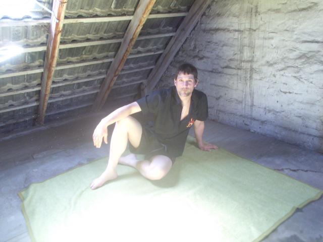 Mario41-hot is sitting on a green mat, barefoot, wearing a black shirt, with his ass and legs spread, in a sunlit, rustic, wooden-shed-like room.