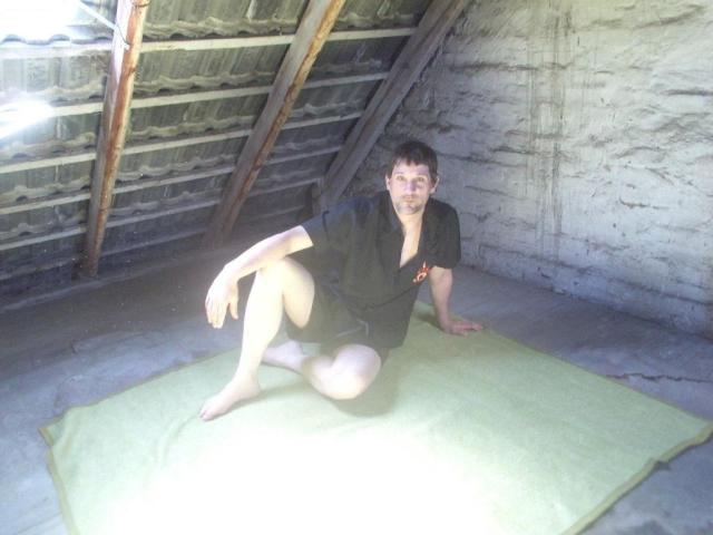 Mario41-hot, a bald dude with short dark hair, is sitting on a green towel in a dirty, wooden-roofed shed, wearing a black shirt and shorts, with a relaxed smile.